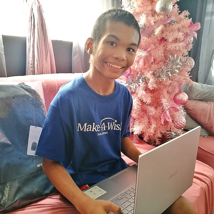 Wish kid Marcus holding his new laptop and sat in a sofa with a Christmas tree behind